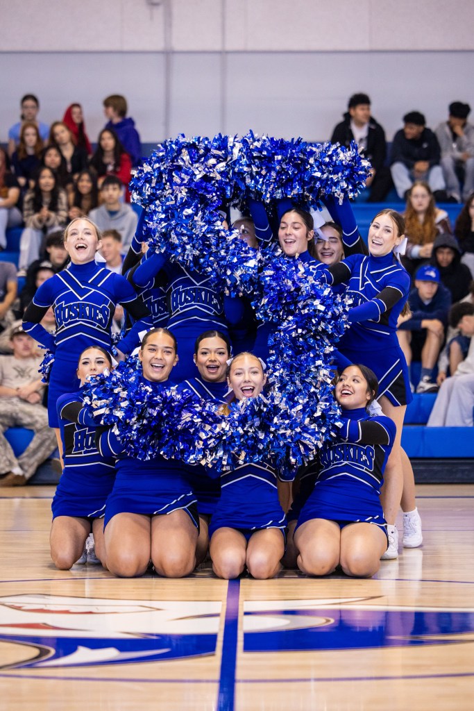 A group of cheerleaders in blue uniforms cheer enthusiastically while holding large blue and silver pom-poms. They form the letter 'H' and are surrounded by spectators in a gymnasium.