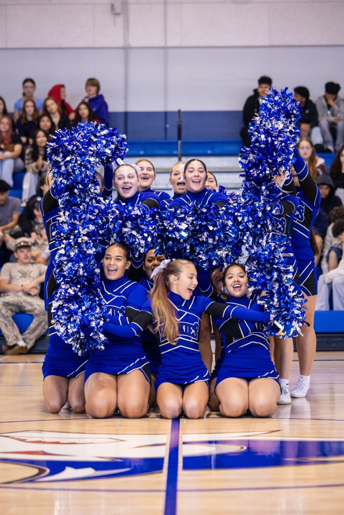 A cheerleading squad performing a formation on a gymnasium floor, smiling and holding blue pom-poms, with a supportive audience in the background.