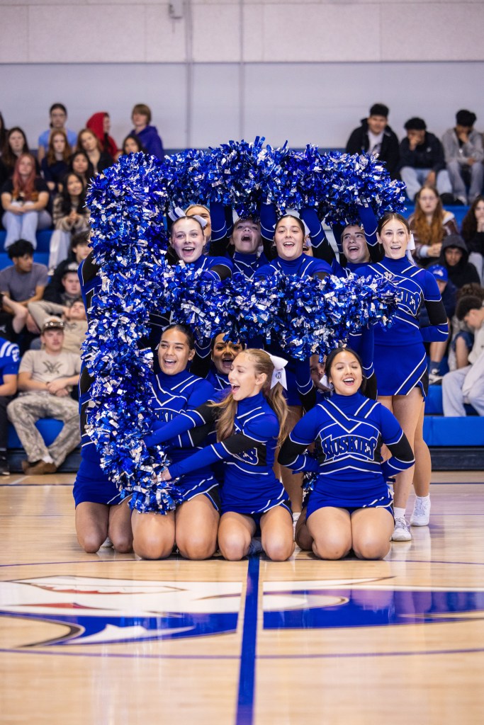 A cheerleading squad forming a large letter with pom-poms during a performance in a gymnasium, surrounded by spectators.