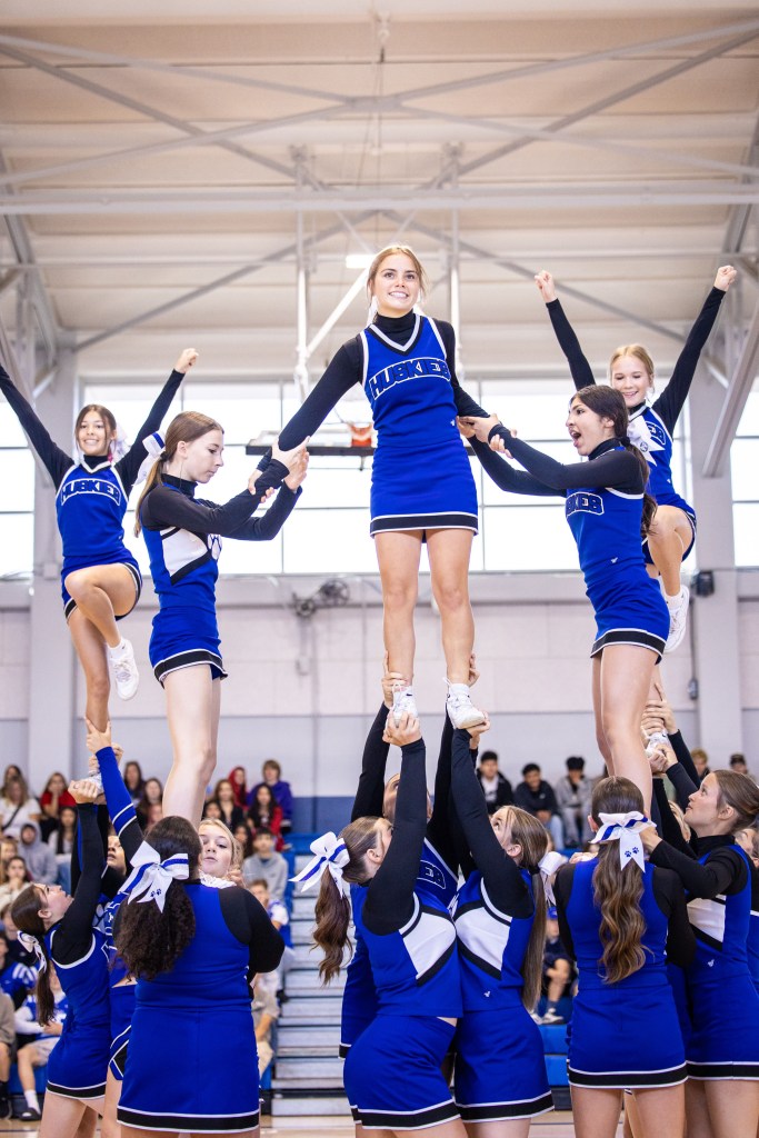 A group of cheerleaders performing a pyramidal stunt, with one cheerleader elevated above others, all wearing blue and black uniforms.