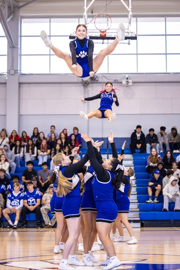 Cheerleaders performing a stunt, with one member in a high jump and another being lifted, in a gymnasium filled with spectators.