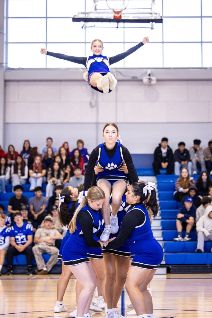 A cheerleading team performs a stunt in a gymnasium, with one cheerleader in the air above two others who are supporting her. The audience in the background observes the performance.