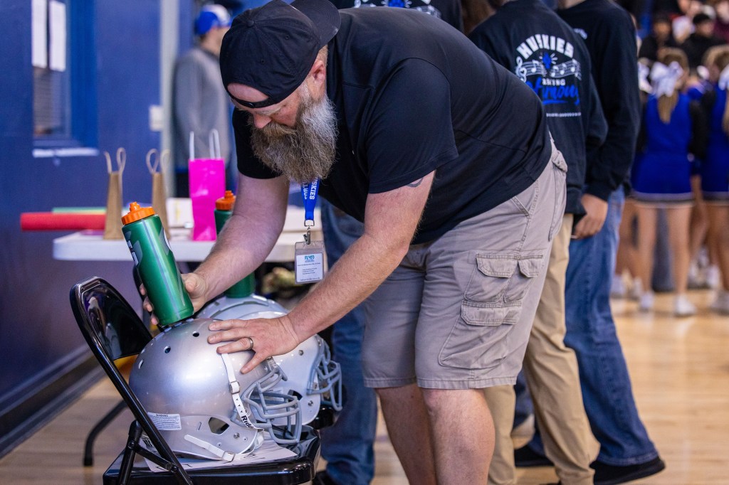 A man with a beard in a black shirt is placing a green sports water bottle on a silver football helmet while standing beside a black folding chair in a gymnasium.