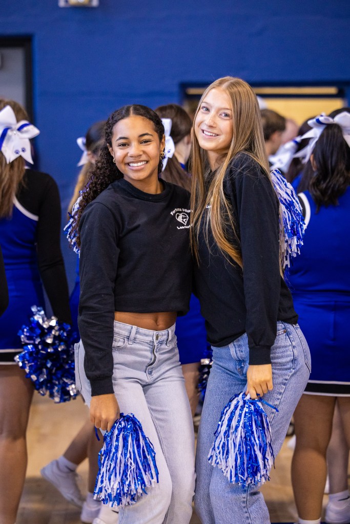 Two cheerleaders smiling together, wearing black sweatshirts and holding blue and white pom-poms, with cheer squad members in the background.