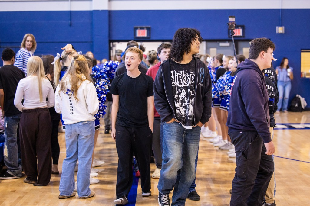 A group of students standing in a gymnasium, with cheerleaders in blue and white uniforms in the background. Some students are engaging with each other, while others are looking in different directions.