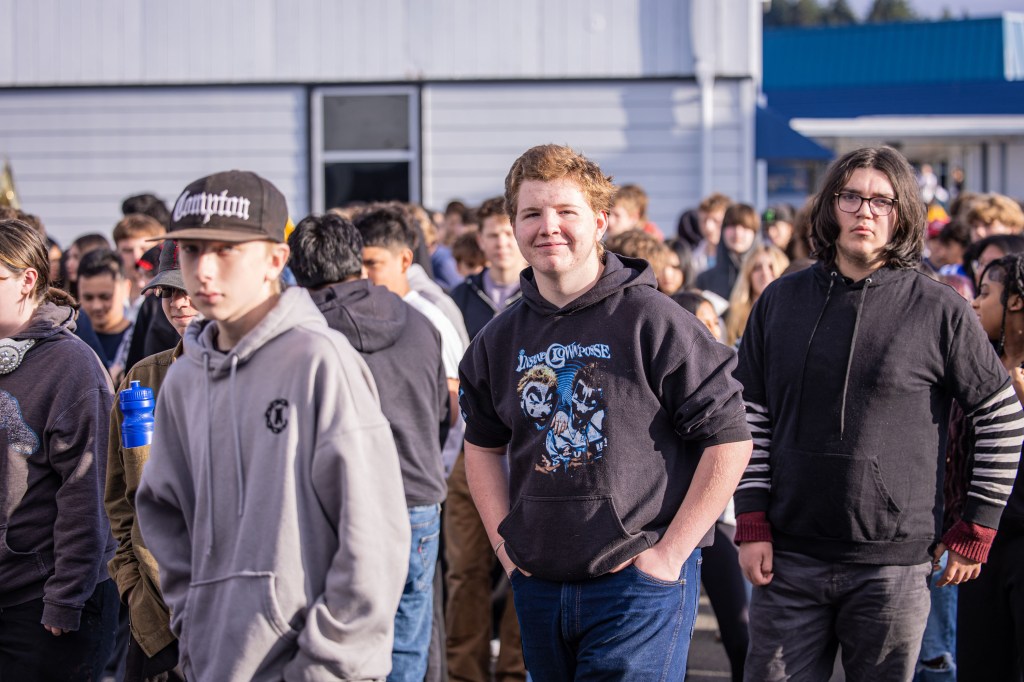 A group of teenagers standing outdoors, some wearing hoodies and casual clothing, with one individual smiling in the foreground.
