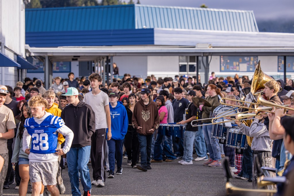A large group of students walking outside a school building, some wearing sports jerseys, while a band plays instruments in the foreground.