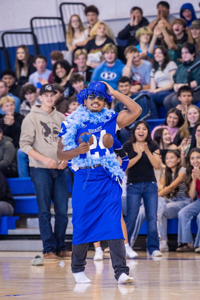 A student in a blue outfit adorned with a feather boa and sunglasses is performing on a gymnasium floor, while a cheering crowd is visible in the background.
