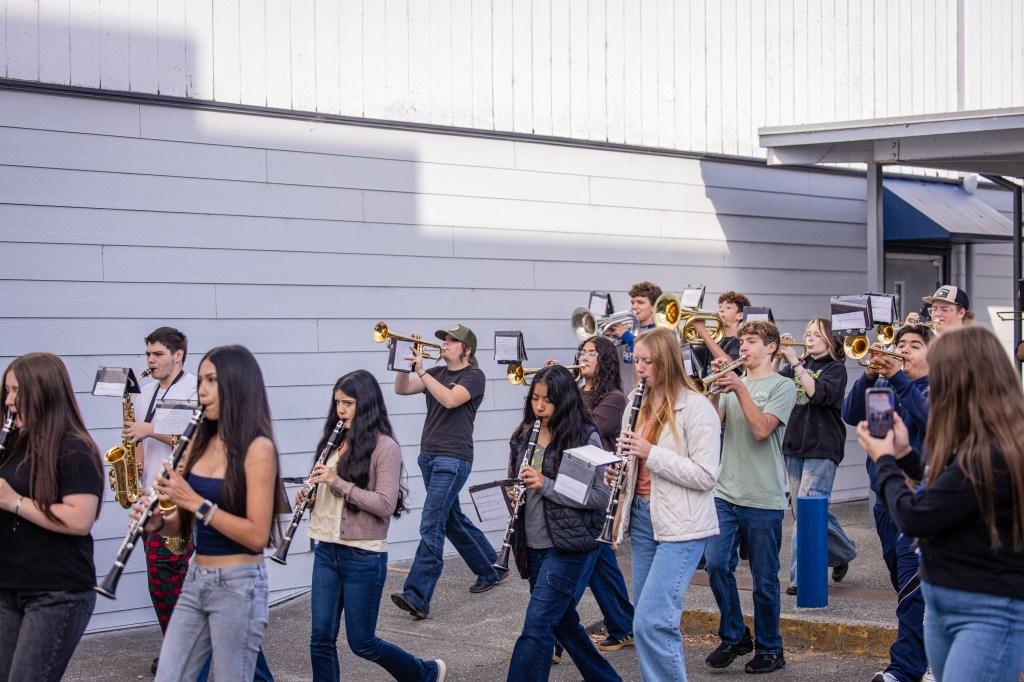 A group of high school students marching in a parade, playing various musical instruments including trumpets and clarinets, with sheet music stands in front of them.