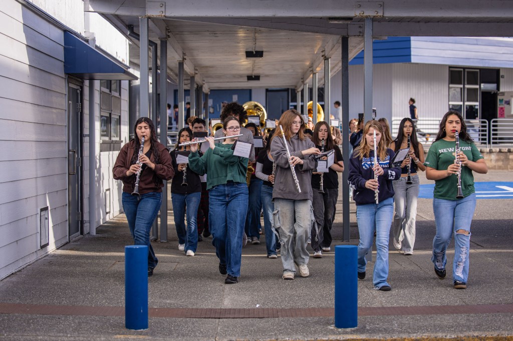 A group of students playing musical instruments while marching down a walkway, with some holding sheet music.