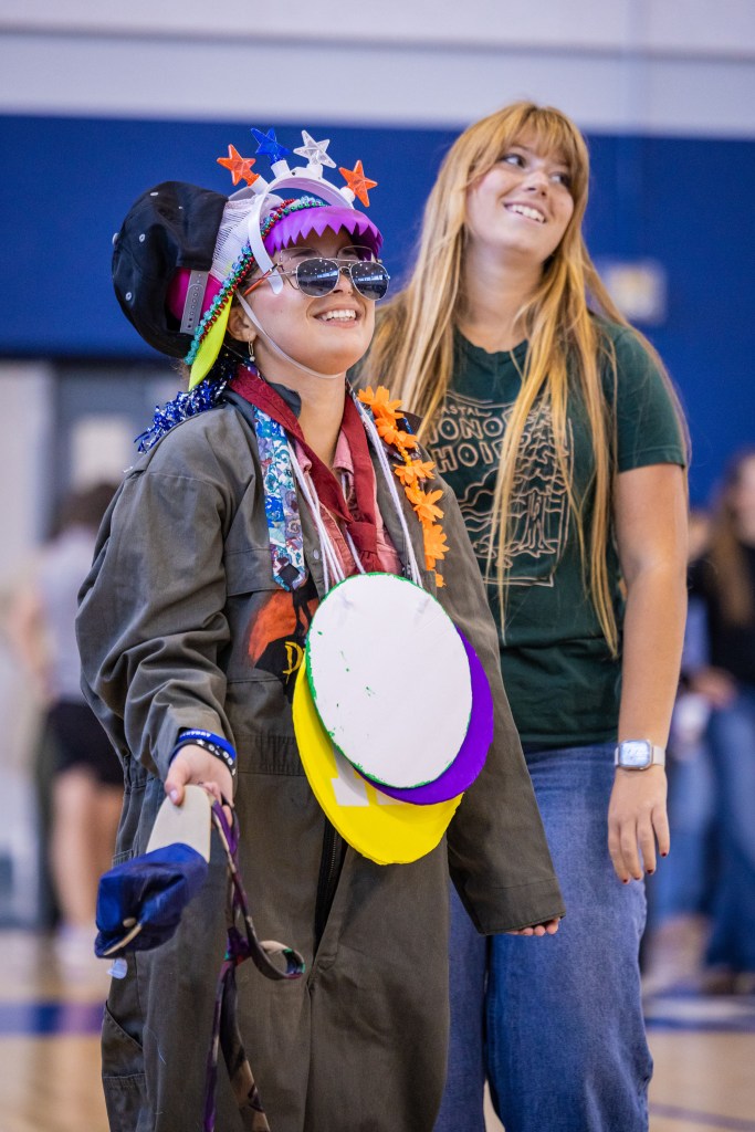Two individuals smiling and posing in a gymnasium. One is wearing a colorful, decorated outfit with sunglasses and a cap, while the other wears a casual t-shirt and jeans.