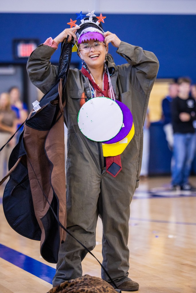 A person dressed in a colorful costume with various decorative elements, including a quirky hat, large circular props, and a playful expression, standing in a gymnasium.