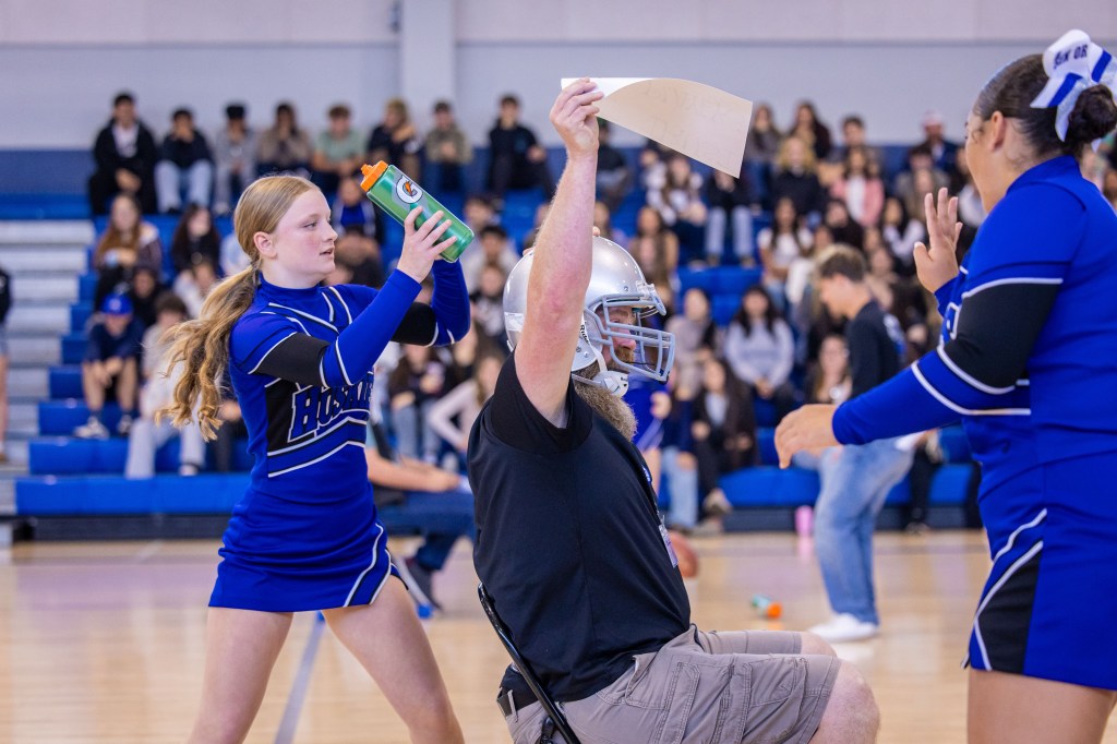 A cheerleader in a blue uniform is holding a water bottle and looking toward a man wearing a football helmet who is sitting with a piece of paper raised in the air. Another cheerleader in a blue uniform is standing beside him. The background shows a gym filled with students watching the event.