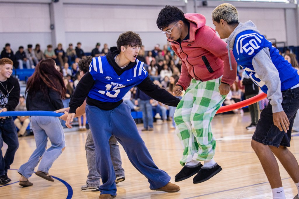 Students participating in a lively school event, jumping over a pool noodle held by classmates, with an audience in the background.