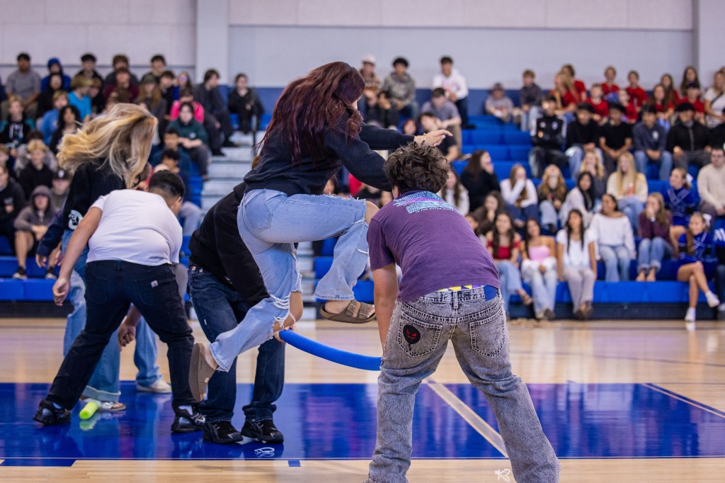 Students participating in a fun activity, jumping over a blue hoop during a school event, with a cheering audience in the background.