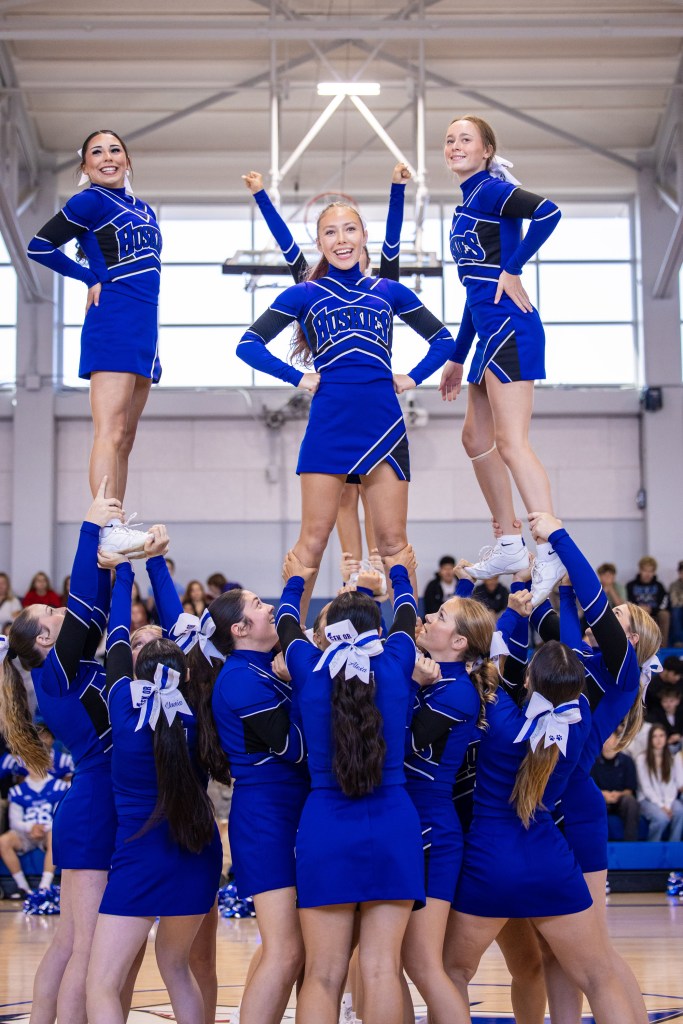 A cheerleading squad performing a stunt in a gymnasium, with one cheerleader elevated on top of a pyramid formed by her team.