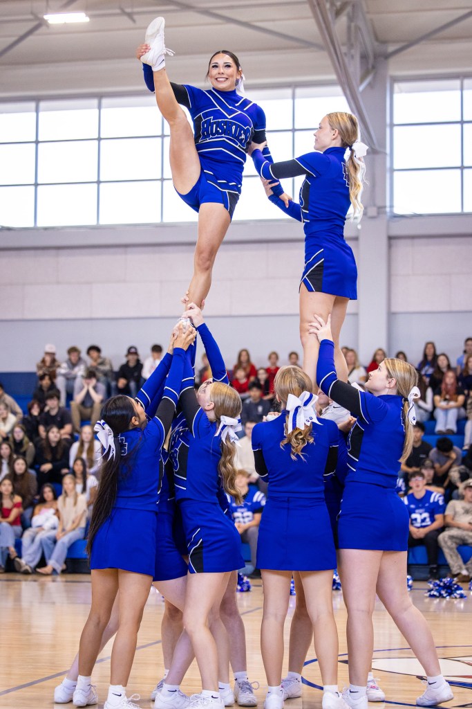 A cheerleading team performs a stunt with one cheerleader elevated on top, displaying flexibility and athleticism, while a supportive group stabilizes her at the bottom, all dressed in blue uniforms.