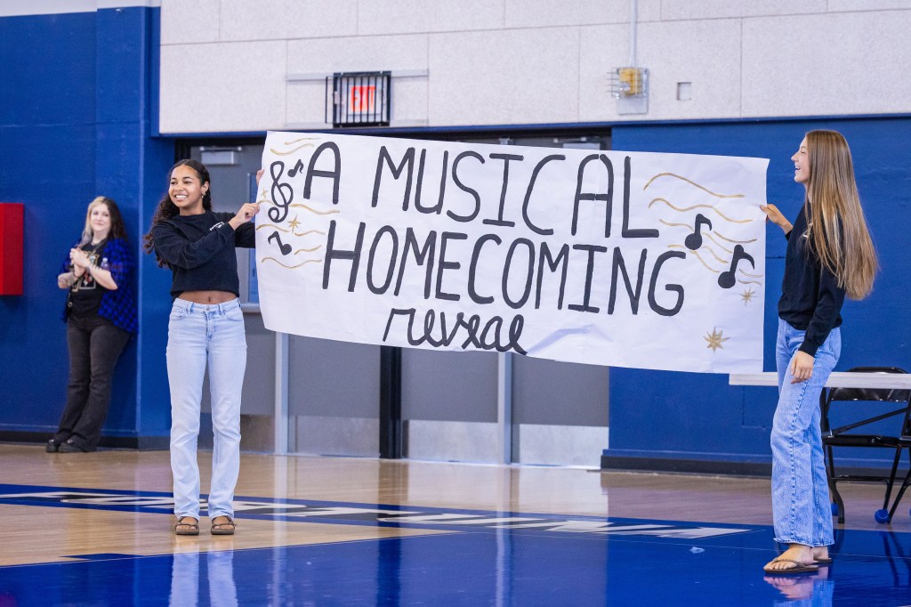 Two students holding a large banner that reads 'A Musical Homecoming Reveal' in a gymnasium setting, with one student smiling and the other gesturing. A third person is seen clapping in the background.