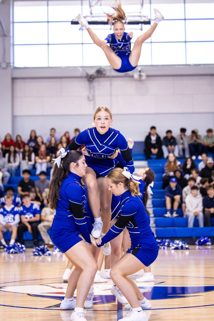 A cheerleading stunt featuring a group of four cheerleaders performing a pyramid, with one cheerleader soaring in the air above them, all dressed in blue and white uniforms.