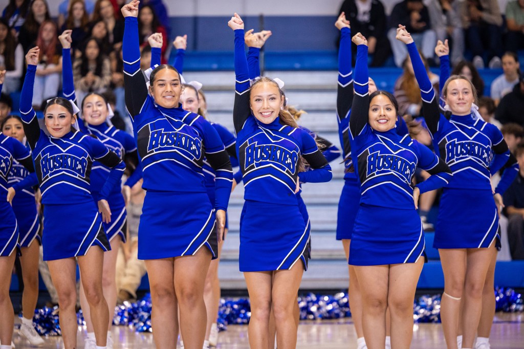 A group of cheerleaders wearing blue and black uniforms, with raised hands and smiling, performing during a school event.