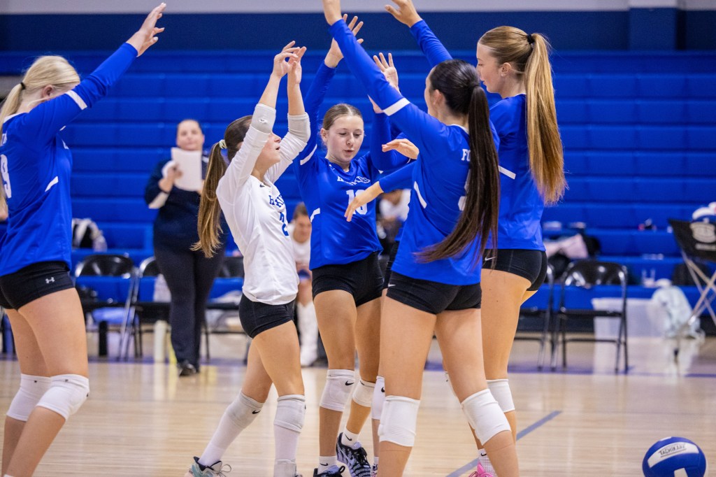 A group of young volleyball players celebrating on the court, wearing blue jerseys and black shorts, with arms raised in excitement.