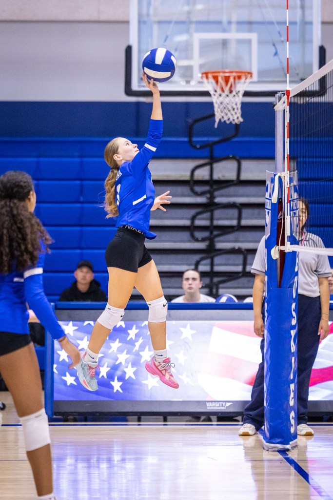 A volleyball player jumps to serve the ball at a sports event, with spectators in the background and an American flag displayed on a digital screen.