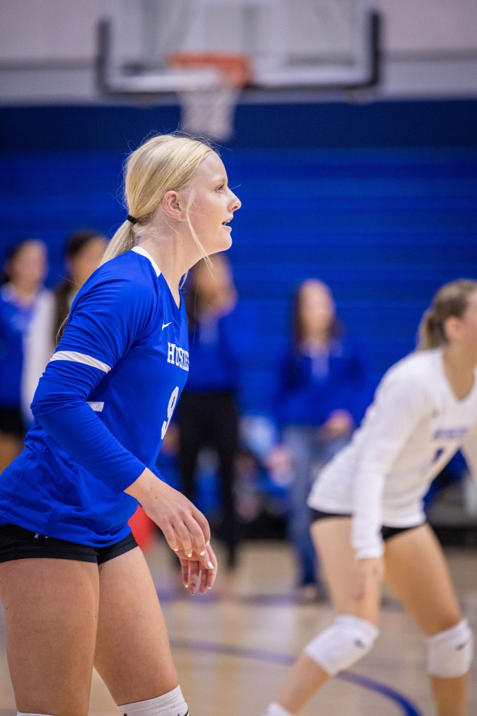 A blonde female volleyball player in a blue jersey prepares for a play during a match, with teammates visible in the background.