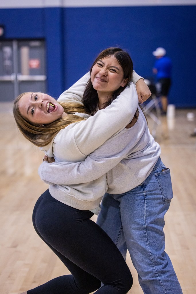 Two young women posing playfully while hugging each other, showcasing joyful expressions in a gym setting.