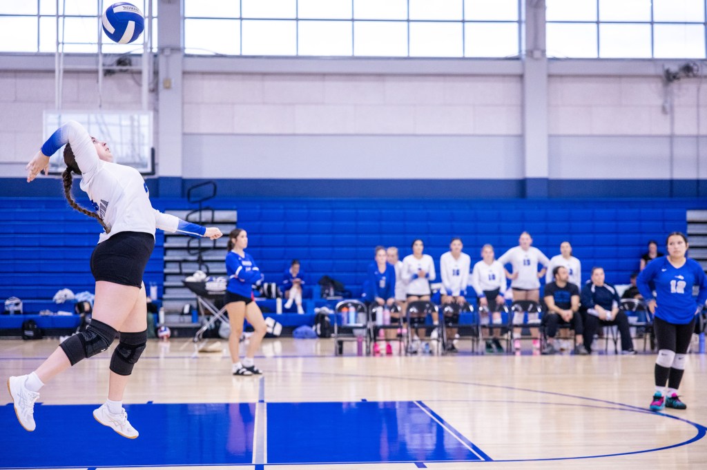 A volleyball player serves the ball during a match, with teammates and coaches watching in the background. The player is in mid-action, preparing to hit the ball, while other players are positioned on the court.