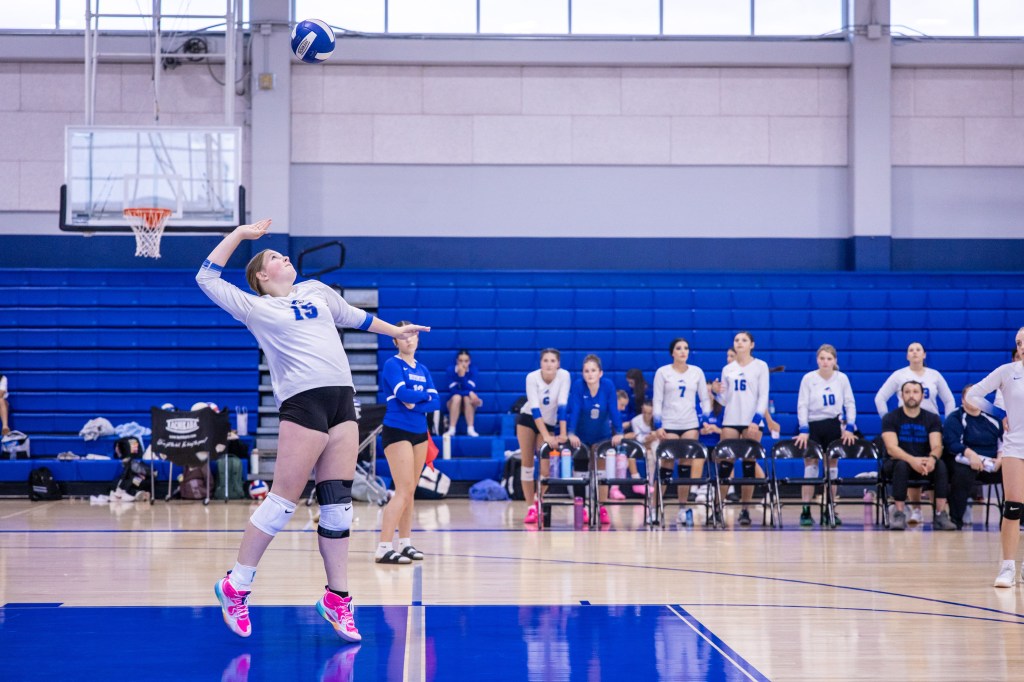 A volleyball player in a gray jersey serving the ball during a match, with teammates in the background sitting on benches.
