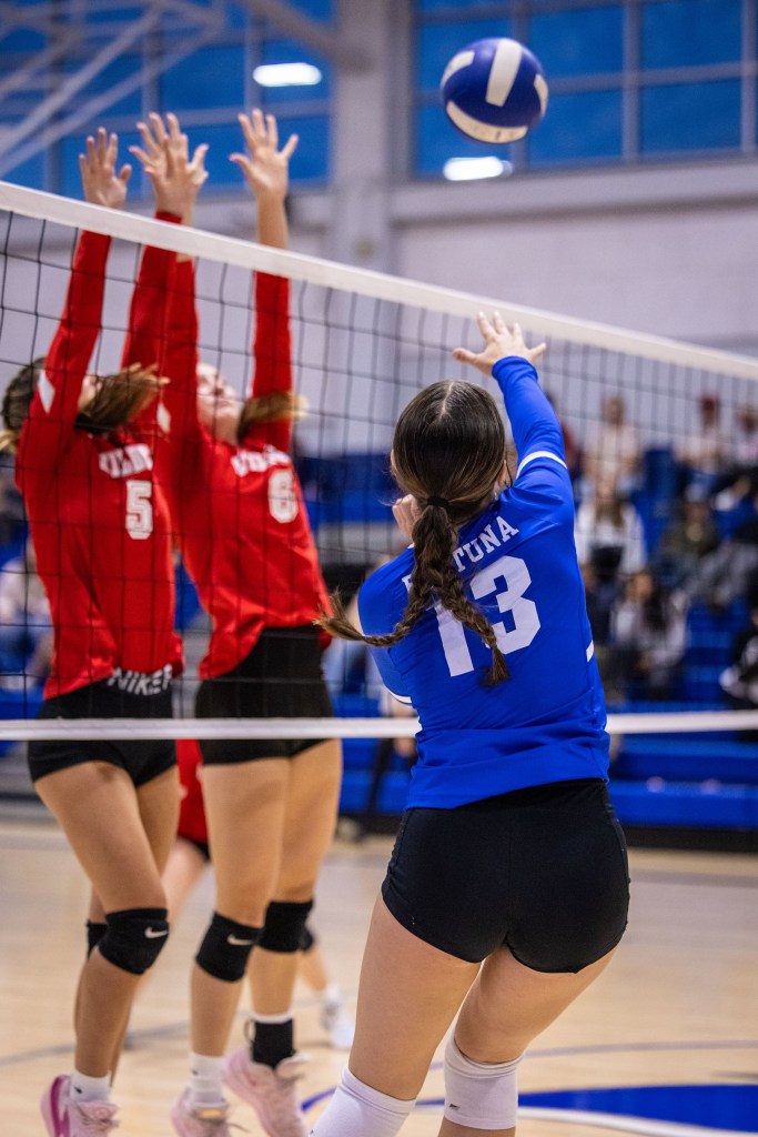 A volleyball match in action, featuring a player in a blue jersey (number 13) attempting to hit the ball over the net, while two defenders in red jerseys jump to block the shot.
