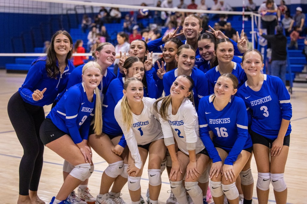 A group of female volleyball players posing together in blue and white uniforms, smiling and showing peace signs, with a volleyball court in the background.