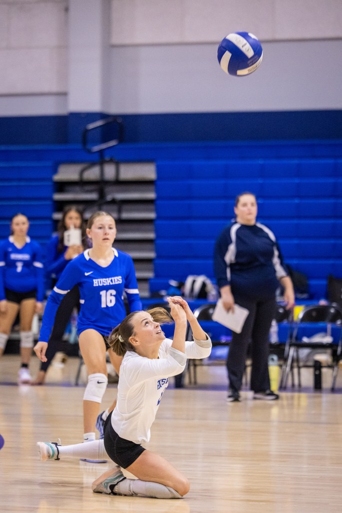 A volleyball player in a white jersey is diving to the floor to receive a volleyball during a game, with teammates and a coach in the background.
