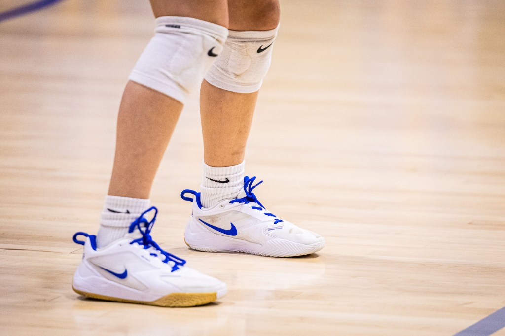 Close-up of a person's feet wearing white Nike volleyball shoes with blue laces and knee pads, standing on a wooden court.