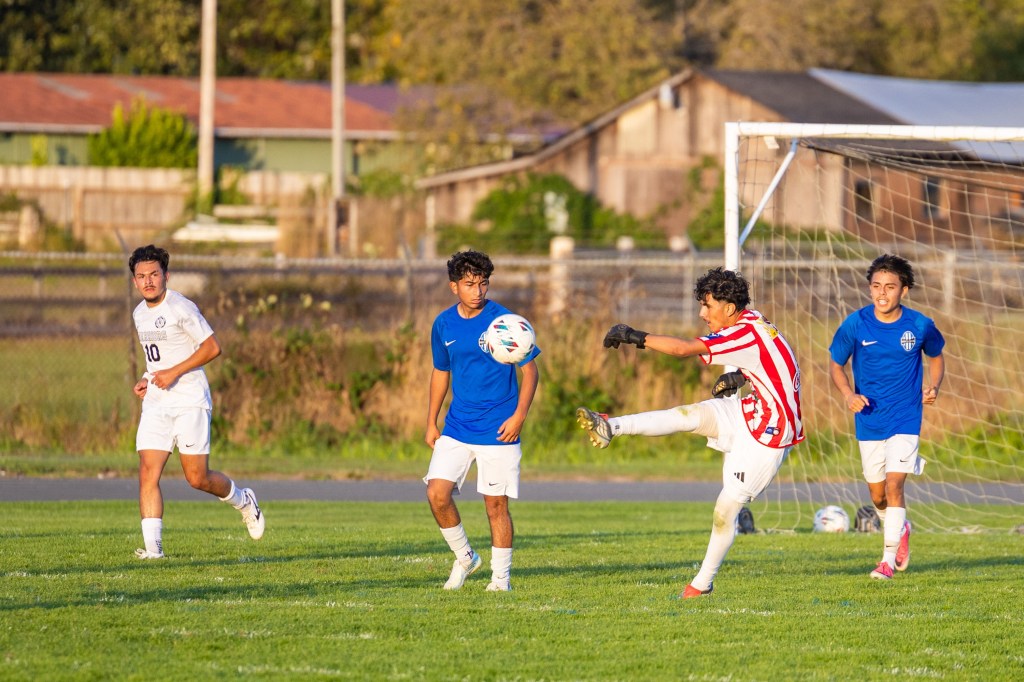 A soccer match in progress with players wearing blue and white uniforms on a grassy field. One player in striped red and white kicks the ball while two defenders watch.