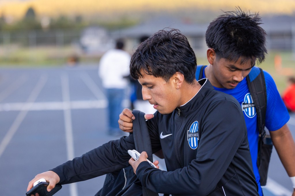 Two young soccer players in team uniforms adjusting their gear on a sports field, with one focusing on a jacket while the other stands nearby.
