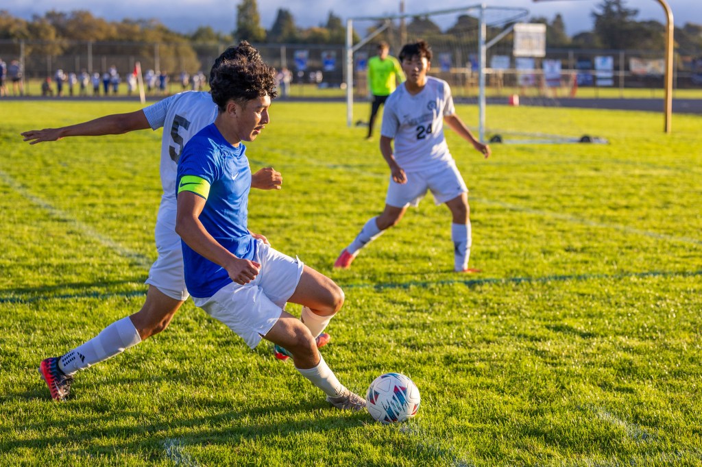 A soccer player in a blue jersey dribbles the ball on a grassy field, while two opponents in white jerseys try to defend. The scene shows dynamic movement in a game setting.