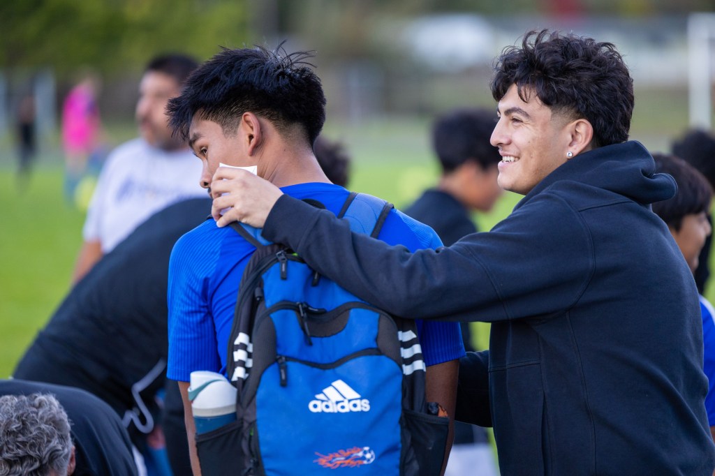 Two young men interacting on a soccer field, one playfully touching the other's shoulder while he is wearing a blue jersey and a backpack.
