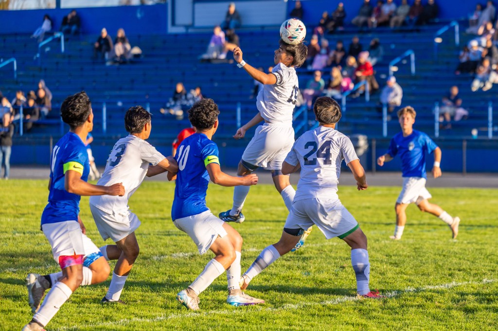 A soccer player in white jumps to head the ball, surrounded by teammates in blue and white uniforms on a grassy field, with spectators in the background.