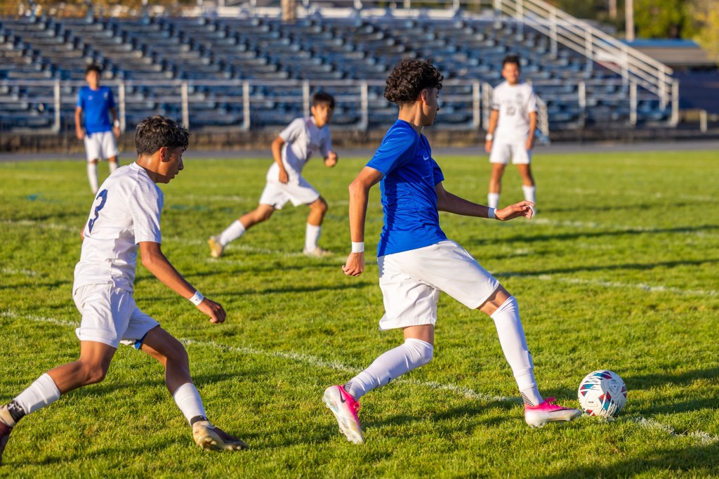 A soccer player in a blue jersey dribbles the ball on a grassy field, while another player in a white jersey attempts to defend.