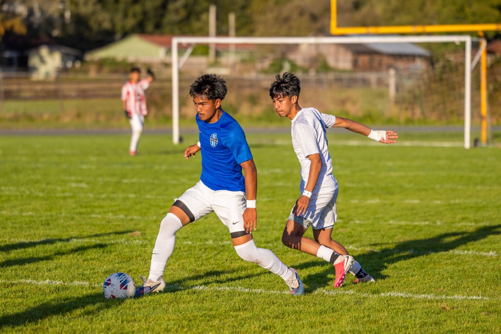 Two young male soccer players compete on a grassy field, with one in a blue jersey dribbling the ball while the other in a white jersey attempts to defend.