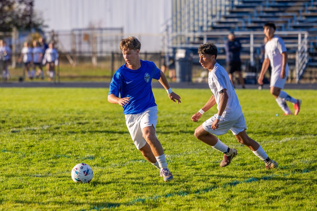 Two high school soccer players on a grassy field, one in a blue jersey and the other in a white jersey, chasing after a colorful soccer ball.