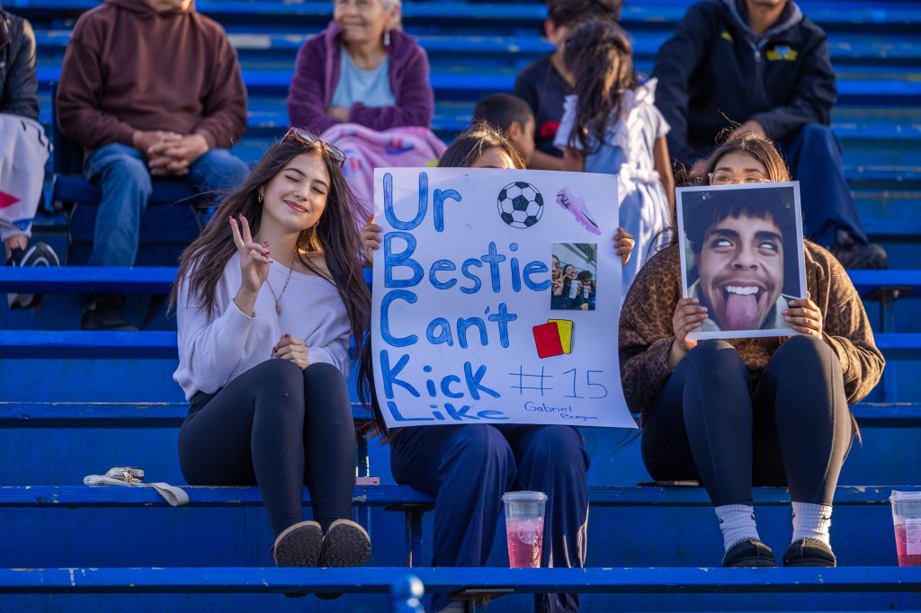 Two young women sitting in bleachers at a sporting event, one holding a sign that reads 'Ur Bestie Cant Kick Like #15' with soccer-themed decorations, while the other holds a photo of a person making a funny face.