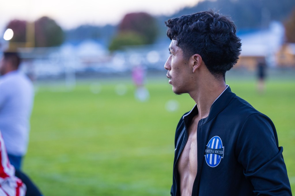 A young soccer player wearing a black jacket with a blue logo standing on a grassy field, looking into the distance.