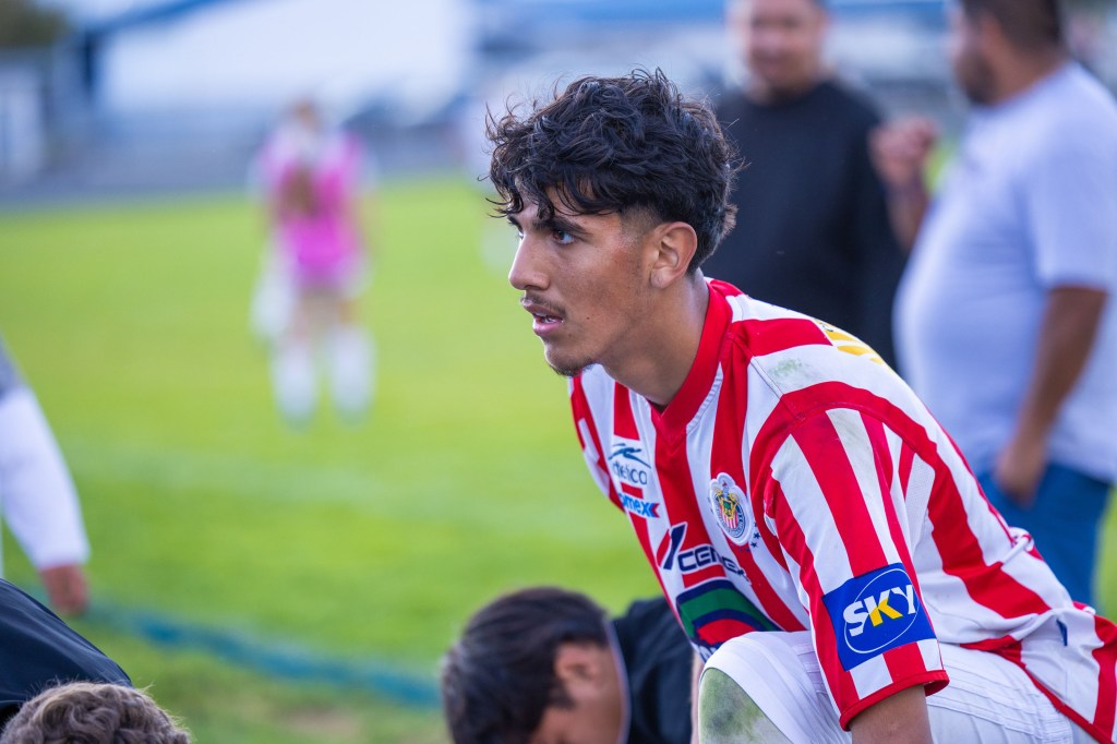A young soccer player wearing a red and white striped jersey sits on the field, looking thoughtfully at the ground, while teammates and spectators are blurred in the background.