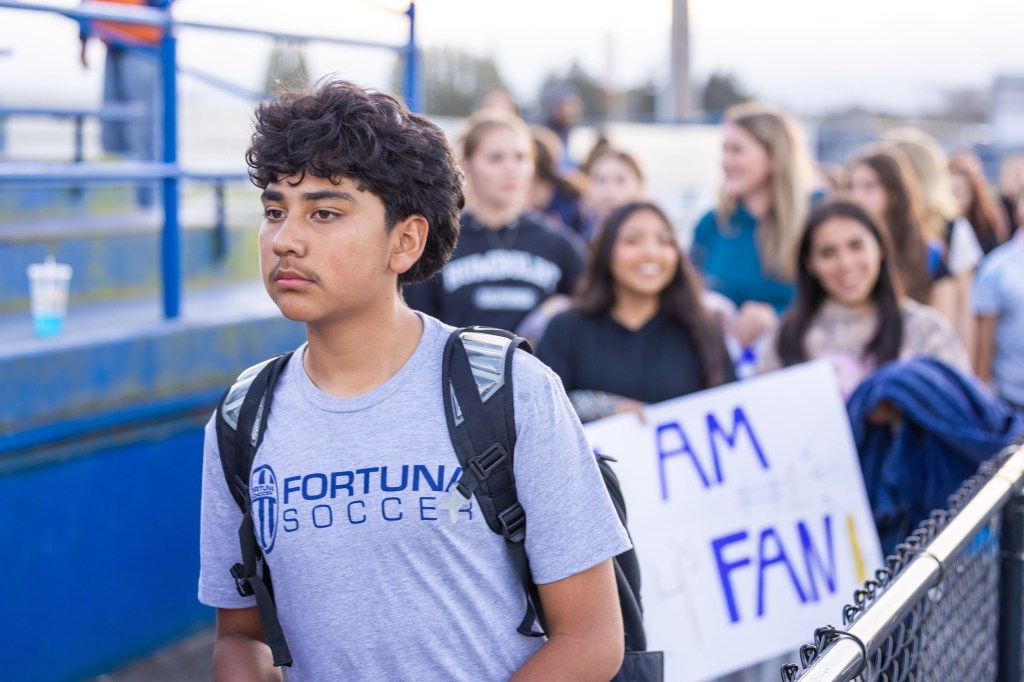 A young boy in a gray Fortuna Soccer t-shirt walks in front of a group of smiling peers holding up a sign that says 'I AM A FAN'.