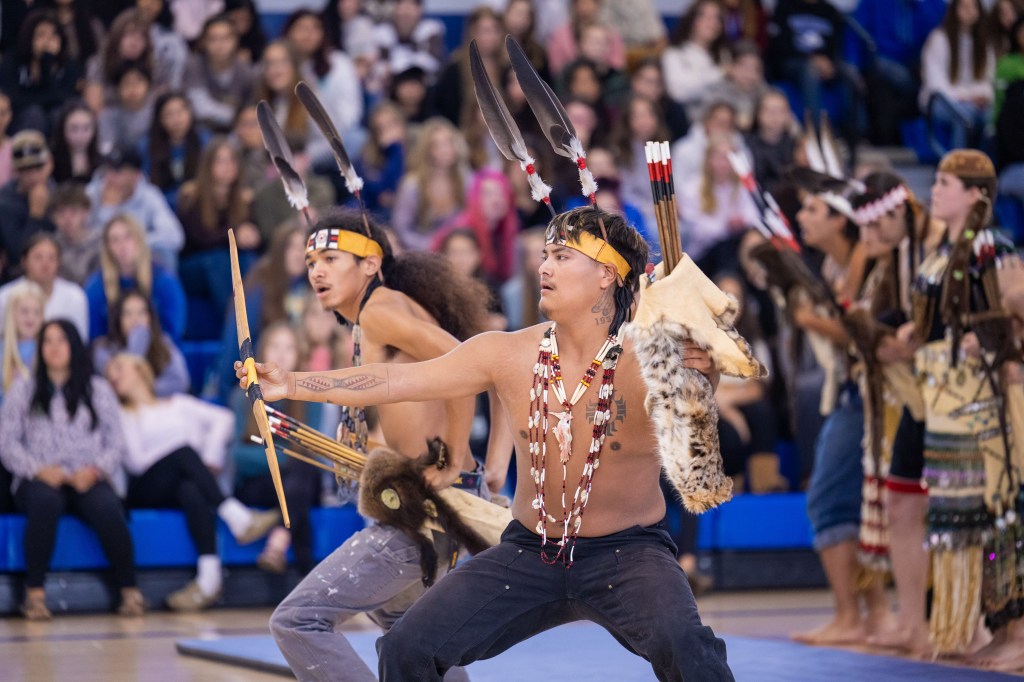 Indigenous dancers performing traditional dance in a gymnasium, wearing colorful regalia and displaying cultural expressions, with an engaged audience in the background.