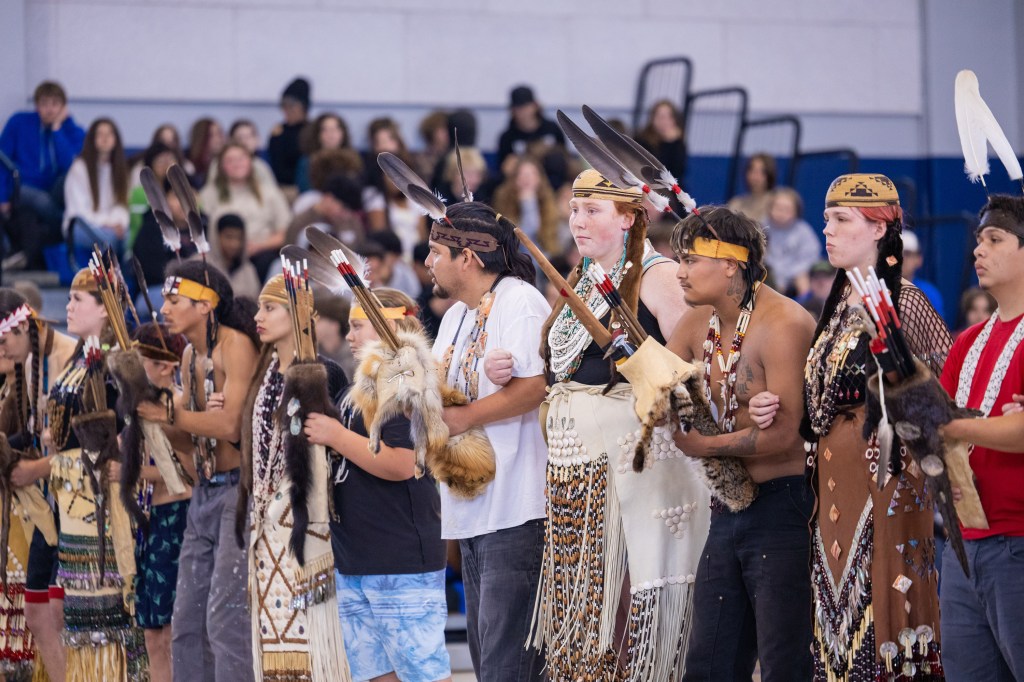 A group of Indigenous dancers in traditional regalia, holding feathered staffs and standing in formation. They are participating in a cultural event, with an audience visible in the background.