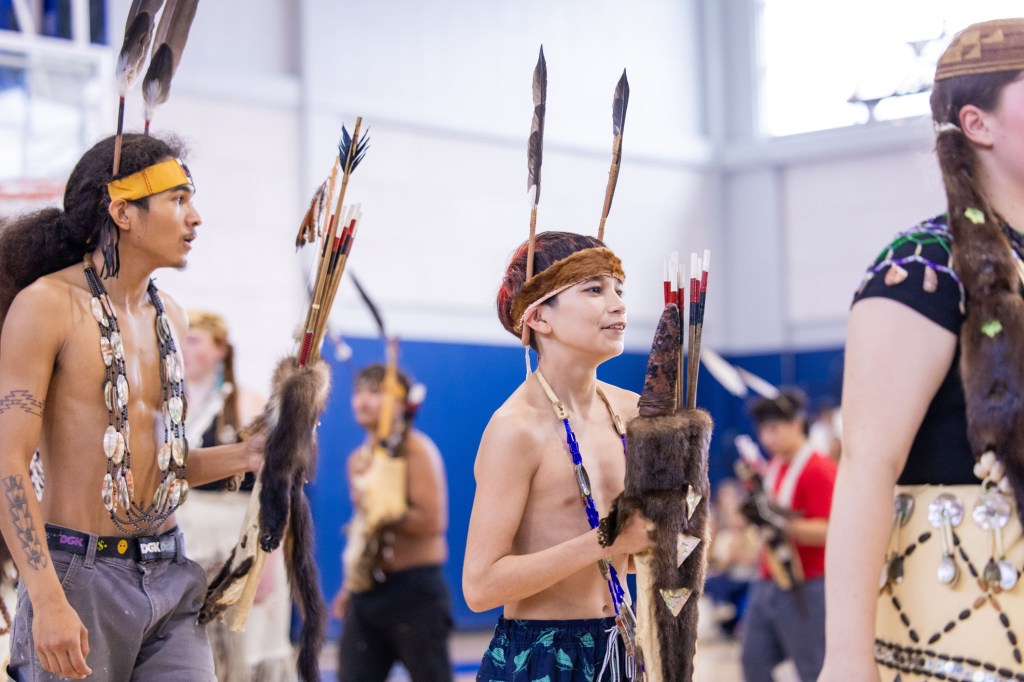 A group of young individuals in traditional attire participating in a cultural dance or event, holding decorated staffs with feathers. The setting appears to be an indoor gym, capturing a moment of celebration and ethnicity.