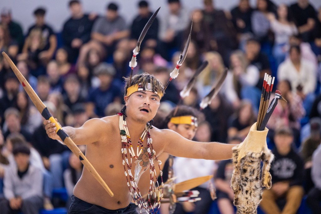 A performer in traditional attire joyfully dancing with a spear and a feathered headdress, surrounded by an audience.